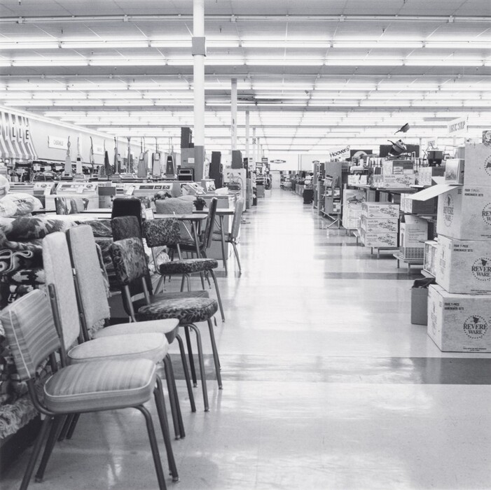 This is a photograph of a large department store interior. The image features a wide aisle with various types of household items on display. On the left side of the aisle, there are rows of chairs with different upholstery styles lined up, while on the right side, stacks of boxes can be seen, labeled "Revere Ware." The store has a high ceiling covered with fluorescent lights providing illumination. Shelving units and multiple aisles are visible in the background, extending into the distance.