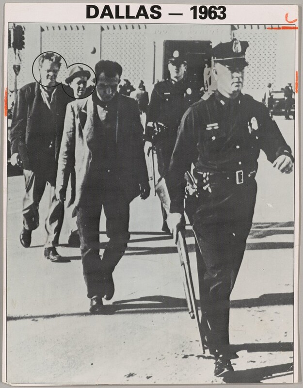 Three men in coats and slacks walk in a line behind a man in a police uniform in this vertical black and white photograph. Another police officer stands or walks near the back of the group. They all are cleanshaven and appear to have light skin, though the face of the man directly behind the police officer at the front is in deep shadow. The officers wear dark uniforms with light-colored badges on the chests, and caps with narrow, curved brims. The officer at the front of the group takes up the rightmost third of the photograph. One arm swings back as he strides forward, and he holds a rifle down by his other side. A revolver is in a holster on that hip. Two of the three men walking behind the officer look down, and the third man there wears a fedora and looks off to our right. The faces of the second and third man are circled with black. The scene is lit from our left, and the people cast long, dark shadows. They walk away from a traffic light on a pole to our left and a building spanning the background. The light-colored walls to either side of the dark doorway of the building appear to be pierced or textured. People stand among cars in front of the building. Typed text in the margin over the picture reads, “DALLAS – 1963.” Short dashes are drawn with orange crayon on the edges of the long sides of the picture near the upper corners. A curving mark over a horizontal orange dash crop the top right corner.
