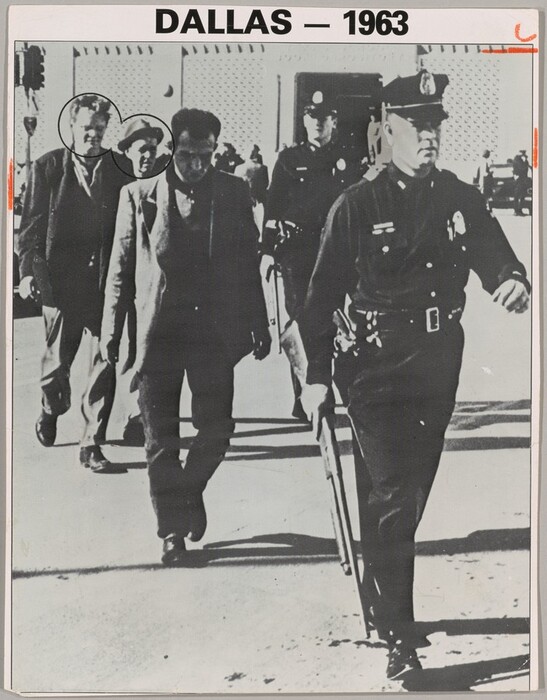 Three men in coats and slacks walk in a line behind a man in a police uniform in this vertical black and white photograph. Another police officer stands or walks near the back of the group. They all are cleanshaven and appear to have light skin, though the face of the man directly behind the police officer at the front is in deep shadow. The officers wear dark uniforms with light-colored badges on the chests, and caps with narrow, curved brims. The officer at the front of the group takes up the rightmost third of the photograph. One arm swings back as he strides forward, and he holds a rifle down by his other side. A revolver is in a holster on that hip. Two of the three men walking behind the officer look down, and the third man there wears a fedora and looks off to our right. The faces of the second and third man are circled with black. The scene is lit from our left, and the people cast long, dark shadows. They walk away from a traffic light on a pole to our left and a building spanning the background. The light-colored walls to either side of the dark doorway of the building appear to be pierced or textured. People stand among cars in front of the building. Typed text in the margin over the picture reads, “DALLAS – 1963.” Short dashes are drawn with orange crayon on the edges of the long sides of the picture near the upper corners. A curving mark over a horizontal orange dash crop the top right corner.