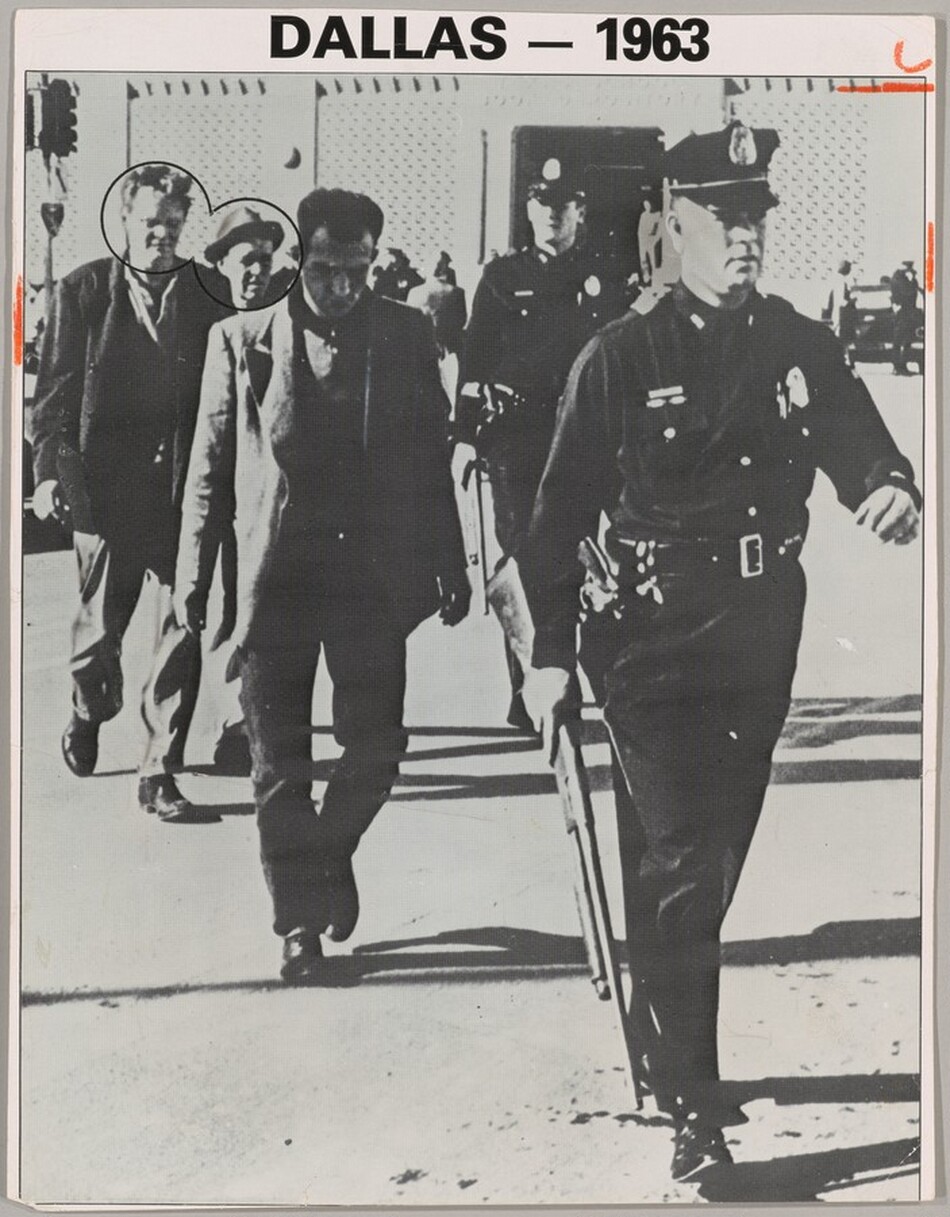 Three men in coats and slacks walk in a line behind a man in a police uniform in this vertical black and white photograph. Another police officer stands or walks near the back of the group. They all are cleanshaven and appear to have light skin, though the face of the man directly behind the police officer at the front is in deep shadow. The officers wear dark uniforms with light-colored badges on the chests, and caps with narrow, curved brims. The officer at the front of the group takes up the rightmost third of the photograph. One arm swings back as he strides forward, and he holds a rifle down by his other side. A revolver is in a holster on that hip. Two of the three men walking behind the officer look down, and the third man there wears a fedora and looks off to our right. The faces of the second and third man are circled with black. The scene is lit from our left, and the people cast long, dark shadows. They walk away from a traffic light on a pole to our left and a building spanning the background. The light-colored walls to either side of the dark doorway of the building appear to be pierced or textured. People stand among cars in front of the building. Typed text in the margin over the picture reads, “DALLAS – 1963.” Short dashes are drawn with orange crayon on the edges of the long sides of the picture near the upper corners. A curving mark over a horizontal orange dash crop the top right corner.