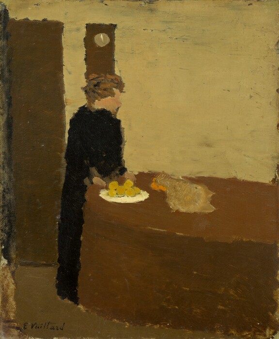 Painted with wide strokes in almost geometric areas of mottled color, a woman wearing a long black dress stands at the side of a table in this stylized, vertical composition. The table stretches two-thirds of the way across the painting from the right edge, and the woman stands at the left side so her lower body is partially behind the table. Her copper-brown hair appears to be pulled up, and her face is painted as a peach-colored profile but without features. Her hands rest on the table near a white plate holding six yellow orbs, presumably fruit. To the right of the plate is a light brown object lined with orange along its left side, but this form is difficult to interpret because the painting is done in such a broad style. Along the left edge of the painting, a dark, pecan-brown door opens next to a rectangular object, probably a clock, on the beige-colored wall behind the woman and table. The floor is a shade between dusky green and brown. The left edge of the cardboard on which this is painted appears streaked, perhaps lightly or incompletely filled in. The artist signed the lower left corner, “E. Vuillard.