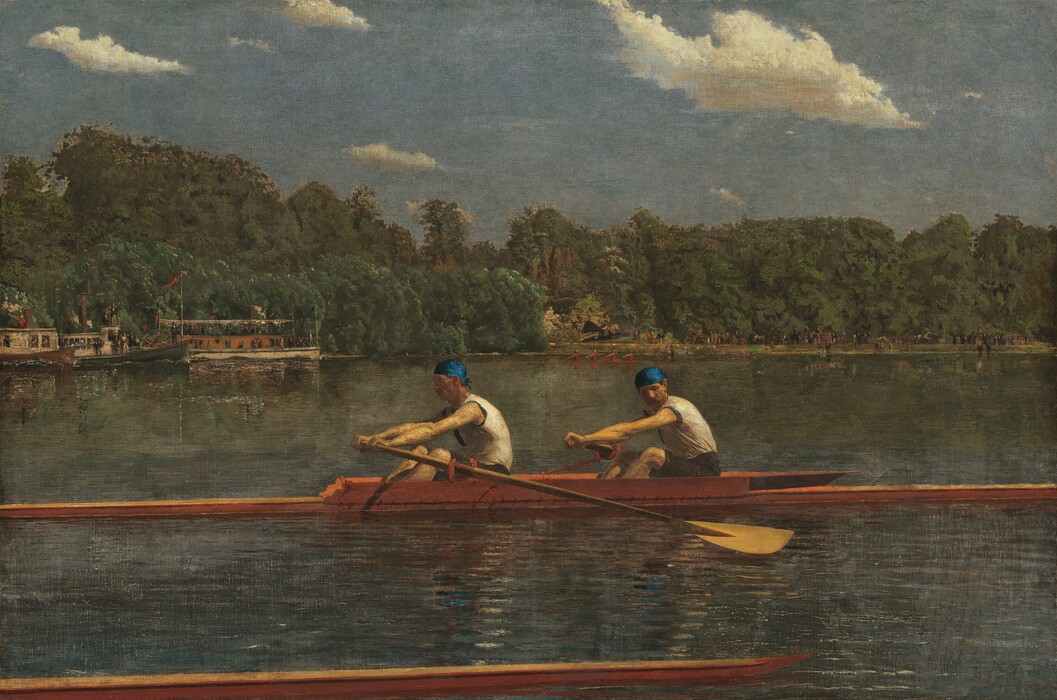Two men with pale skin, wearing white sleeveless shirts and royal-blue caps, row in unison in a long, narrow boat on a placid blue river in this horizontal landscape painting. The low, honey-colored wooden boat extends off both sides of the canvas. Both men face our left as they row to our right. The man in front looks ahead of him, beyond the stern of the boat, and the man to our right, closer to the bow, tucks his chin down to look past his shoulder. Their bare, muscled arms are extended straight as the two oars sweep back. The tip of another boat runs close and parallel to the bottom edge of the composition, spanning the left three-quarters of the painting. The opposite riverbank is lined with a dense forest of pine-green trees. People crowd along the decks of a steamboat and a paddleboat near the riverbank to our left. Another narrow skuller, rowed by four people wearing ruby-red shirts, cuts through the water at the back center of the river. The riverbank beyond is lined with people, painted with strokes of black and white, and minuscule touches of red. The horizon comes halfway up the composition. Cream-white clouds float across a muted, topaz-blue sky above the trees.