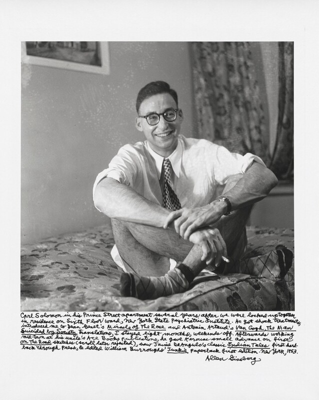 A man sits smiling, cross-legged on a bed with his elbows wrapped around his raised knees in this square black and white photograph. The man’s body is angled slightly to our right, and he looks off, grinning, in that direction. His short dark hair is brushed up and back away from his forehead, and he wears dark, round-framed glasses with thick lenses. The man’s white button-down shirt is rolled up to the elbows, and he wears a tie patterned with dark diamonds against a light background. His hairy legs are bare, and he wears argyle-patterned socks. On one arm, he wears a wristwatch and grips his other wrist with that hand. He holds a nearly burned-down cigarette with that other hand. The bedspread has floral patterns, and curtains are blurry on the far right side of the room. Handwriting below the photograph reads, “Carl Solomon in his Prince Street apartment several years after we were locked up together in residence on Sixth Floor Ward, New York State Psychiatric Institute. He got shock treatments, introduced me to Jean Genet's Miracle of the Rose and Antonin Artaud's Van Gogh, The Man Suicided by Society translations, I stayed eight months, weekends off. Afterwards, working mid-town at his uncle's Ace Books publications, he gave Kerouac small advance on first On the Road sketches (scroll later rejected), saw Jaime DeAngulo's classic Indian Tales first hard-back through press, and edited William Burroughs' Junkie paperback first edition. New York, 1953. Allen Ginsberg.”
