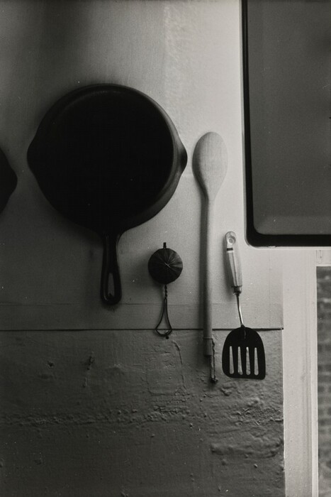 A large cast iron skillet, a wooden spoon, and a slotted spatula hang on a wall, accompanied by a small round object that looks like a kitchen timer. The image is in black and white. The skillet is on the far left, followed by the timer, the wooden spoon in the middle, and the spatula on the right near the edge of the frame. The objects are positioned vertically against a textured wall that shows a slight divide between the upper and lower sections.