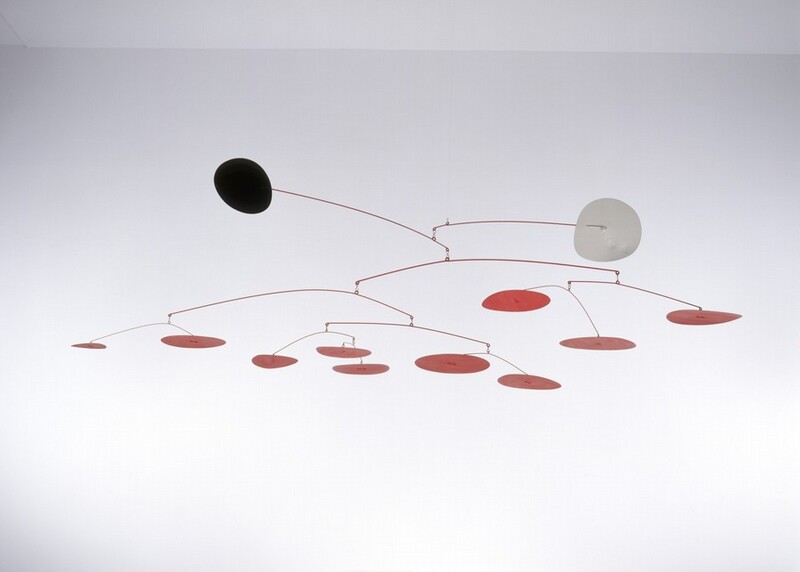 Two disks, one black and one white, and ten smaller red disks hang from eleven red wire branches in this free-floating sculpture, called a mobile. The uppermost branch ends with the white disk jutting out to our right, and the next ends with the black disk, projecting out and slightly up to our left. The red disks cluster in a loose line below, some higher and some drooping down. The mobile is suspended with an unseen filament in a white room in this photograph.