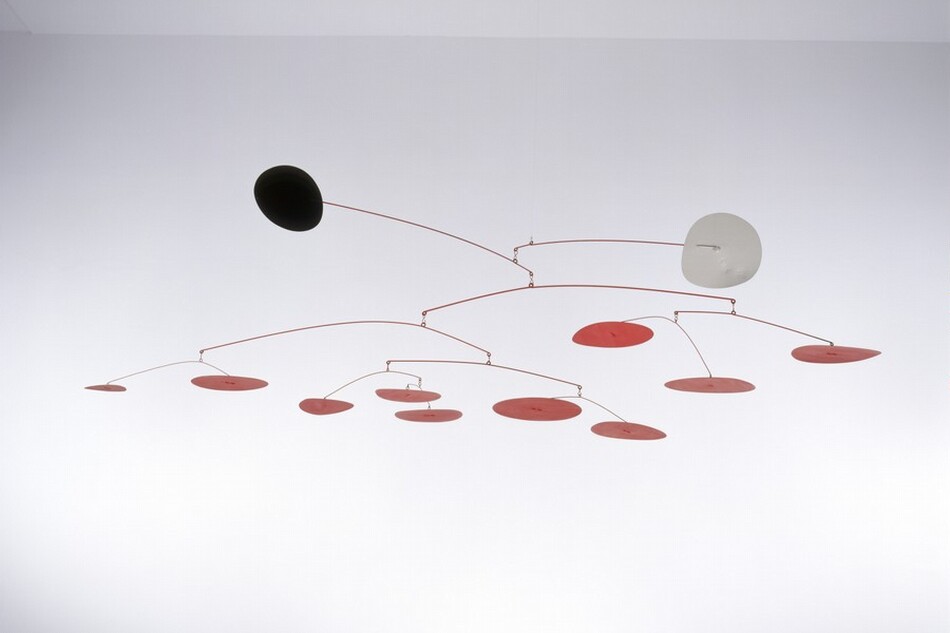 Two disks, one black and one white, and ten smaller red disks hang from eleven red wire branches in this free-floating sculpture, called a mobile. The uppermost branch ends with the white disk jutting out to our right, and the next ends with the black disk, projecting out and slightly up to our left. The red disks cluster in a loose line below, some higher and some drooping down. The mobile is suspended with an unseen filament in a white room in this photograph.