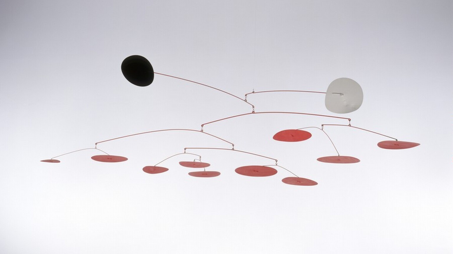 Two disks, one black and one white, and ten smaller red disks hang from eleven red wire branches in this free-floating sculpture, called a mobile. The uppermost branch ends with the white disk jutting out to our right, and the next ends with the black disk, projecting out and slightly up to our left. The red disks cluster in a loose line below, some higher and some drooping down. The mobile is suspended with an unseen filament in a white room in this photograph.
