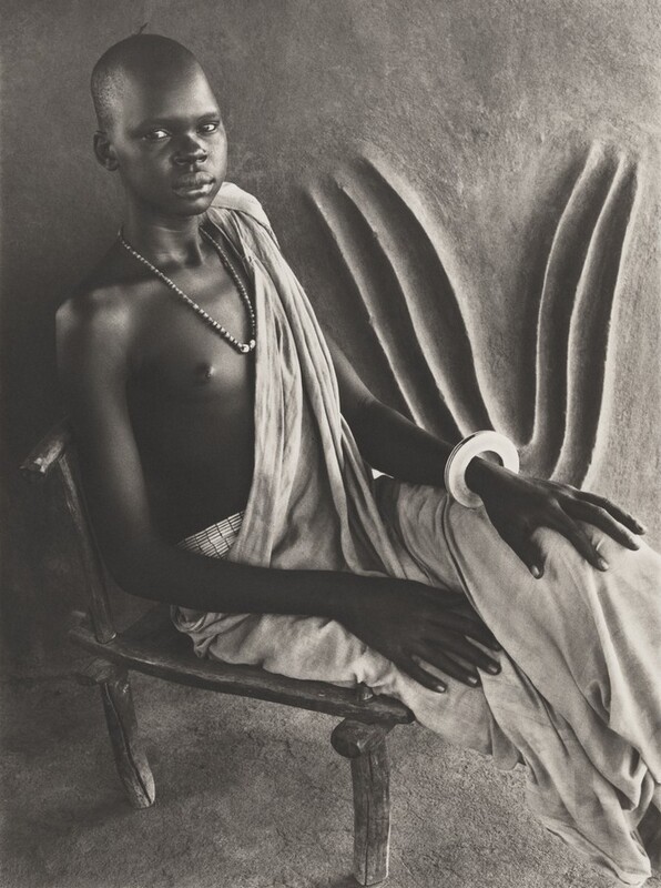 This grayscale photograph shows a person seated on a rustic wooden chair, with their body turned slightly to the right as they look towards us. Their head is shaved. They are wearing a draped cloth garment that covers their lower body and one shoulder, leaving the other shoulder bare. Around their neck is a strand of beads, and they have a large bracelet on one wrist. The background features a plain textured wall with some curved patterns carved in relief.