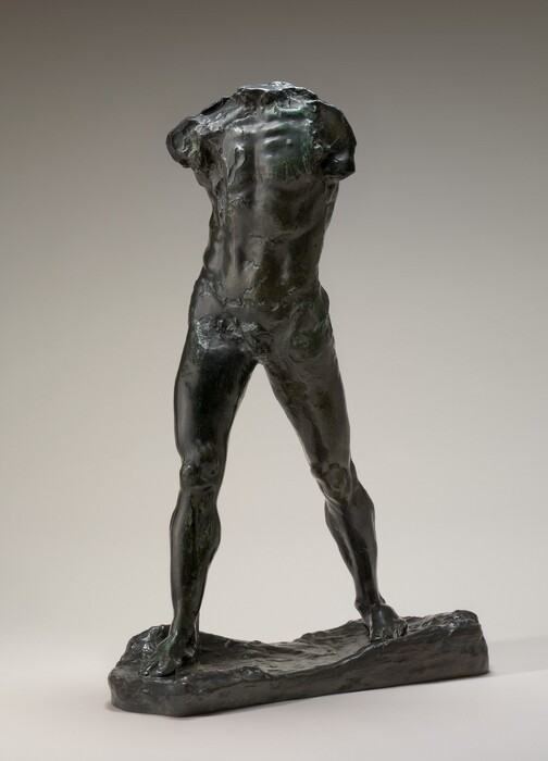 The sculpture depicts a male torso and legs without a head or arms. The body is nude, and its legs are spread apart, standing on an uneven base. The body faces us. It is crafted from a dark brown material, and some areas have a greenish sheen.