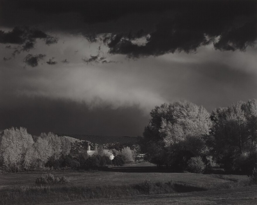 The image shows a landscape with grassy land stretching into the distance. There are trees and a structure resembling a chapel with a bell tower at the horizon. The sky above has dark clouds mixed with lighter patches. The trees are highlighted by light, contrasting with the sky. The ground close to the viewer has textured grass. The image is in black and white, emphasizing light and shadow contrasts.