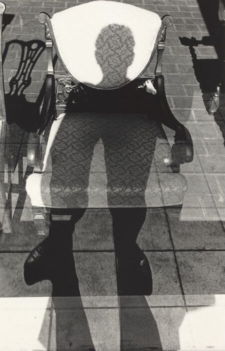 This is a photograph of reflections and shadows on a textured chair and tiled floor. The image shows a figure being reflected on the chair and casting a shadow on the floor. The chair has intricate woodwork and an ornate upholstery pattern. The shadow appears elongated with discernible legs and footwear, aligning with the reflection seen through a glass surface. The tiled floor adds a structured background to the composition. The play of light and shadow creates a juxtaposition of textures and forms in the image.