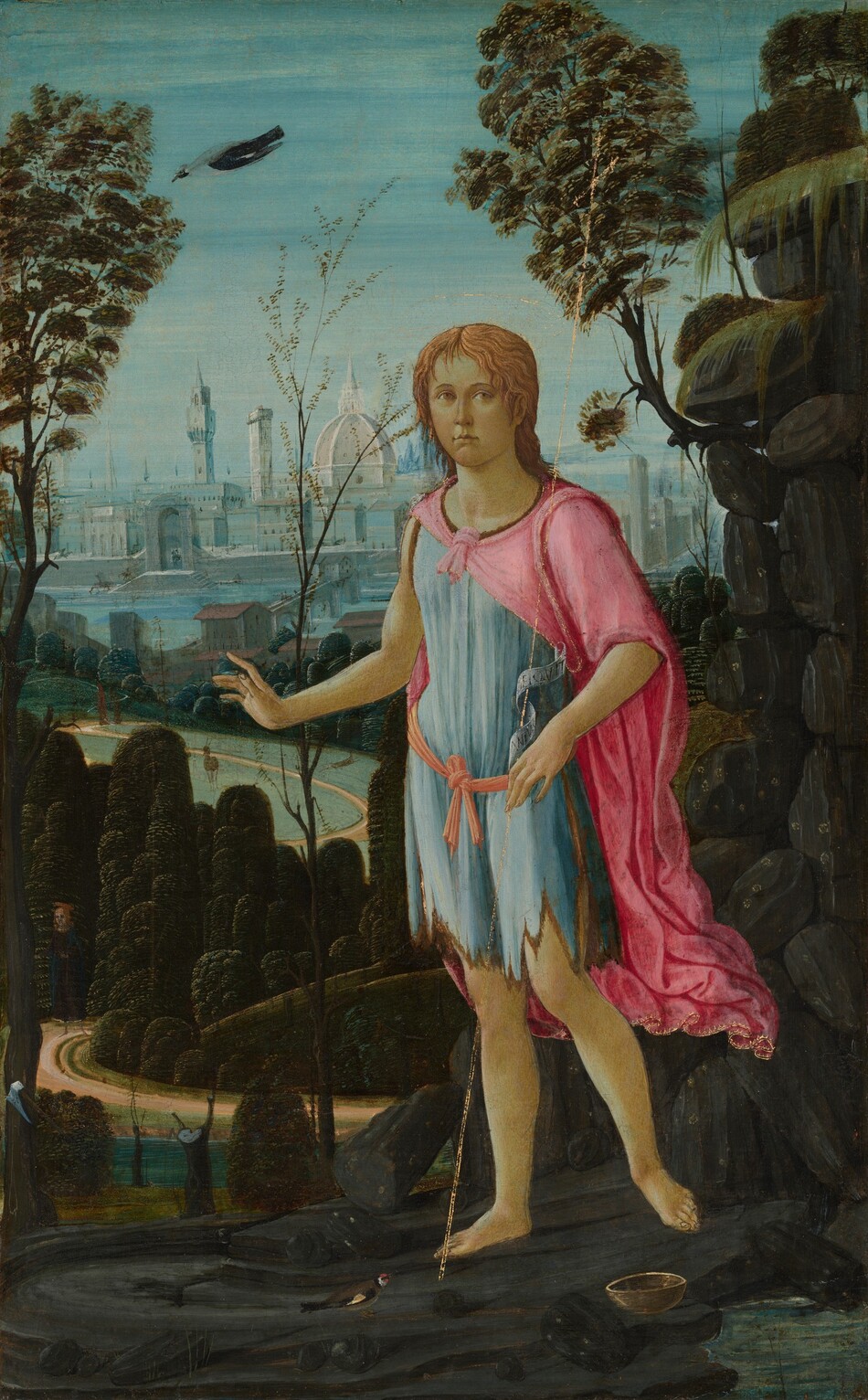 This painting shows a person standing on a hill in rocky terrain. The person has light skin and short yellow hair. They are wearing a light blue tunic with jagged edges, tied at the waist with a pink sash. A pink cloak is draped over their shoulders. There is a small black bird near their feet, and another bird flies overhead. They appear to hold a thin, faded golden cross in one hand that extends above their head. There are hints of gold around their head, suggesting there once may have been a golden halo. The background includes lush greenery, trees, and stacked gray rocks on the right side. In the distance below is a winding path that leads through dark trees to a blue and white cityscape with large white domed buildings. The sky above is blue and cloudless.