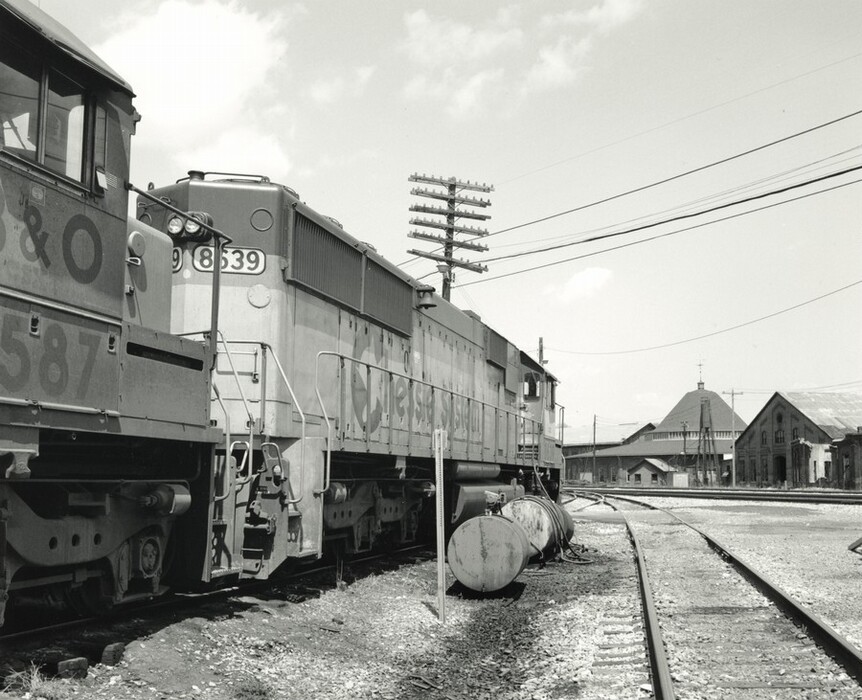 The image shows a train yard with two locomotives in focus. The view is from track level, showcasing the train engines, with one displaying the number 9839. The ground is covered with gravel and tracks, leading towards railroad buildings in the distance. The horizon is partially obscured by structures such as a roundhouse with a conical roof and a rectangular building. The sky is mostly clear with some clouds, creating a bright scene in the black and white photograph. The contrast between the shadows on the trains and the light sky indicates a sunny day. Tall telegraph poles run parallel to the tracks, with overhead wires crisscrossing above the locomotives.