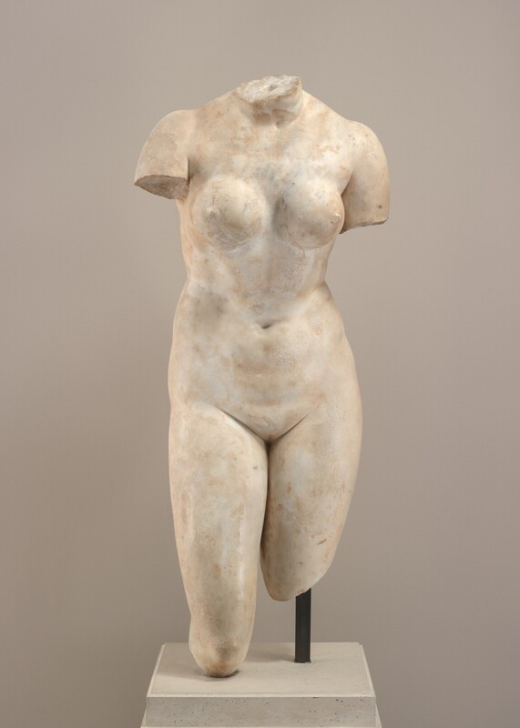 A female torso from the neck to the shoulders and knees is carved from white marble in this freestanding sculpture. The body is square toward us in this photograph. The neck is jagged where the head has broken off, and the arms are missing below the shoulders. The leg on our right is broken off above the knee and the other below the knee. Firm, round breasts are above a soft belly and smooth groin. Some areas of the surface are smooth and others are rough, and the marble has darkened to tan in places. The sculpture is supported by a metal rod on our right, and the work rests on a square base. The wall behind is light gray.