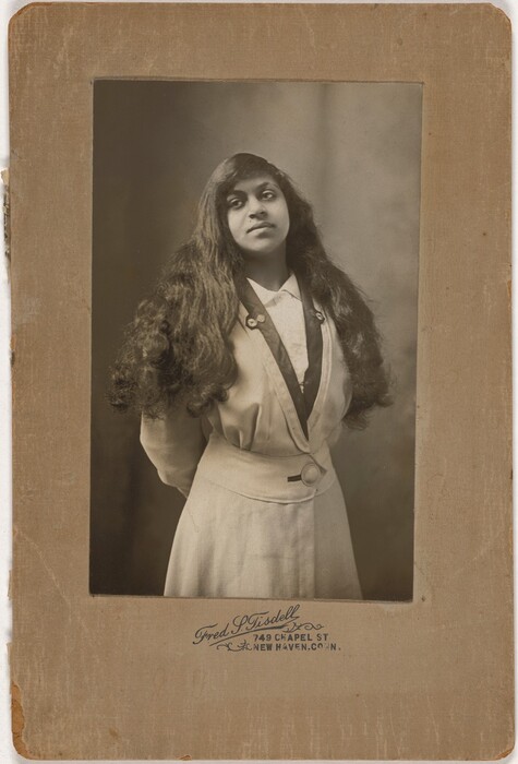 The image depicts a woman standing from the waist up. She has her hands behind her back and is wearing a light-colored blouse with a dark collar and two buttons. The woman has long, dark hair flowing past her shoulders in soft waves, and she appears to have a serious expression with expressive eyes. She is wearing a skirt or dress with a defined waist, secured by a belt with a circular buckle. The background is plain and blurred, and the photograph is mounted on a cardboard frame with an inscription indicating the photographer and location in New Haven, Connecticut.