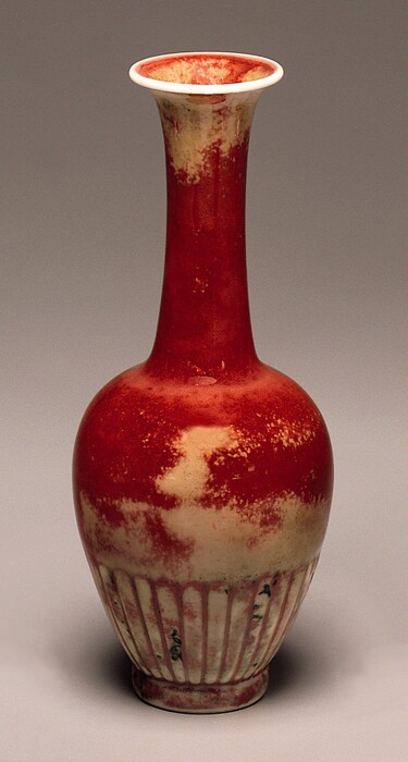 This sculpture is a vase with a long neck that flares out at the rim. The center of the vase is primarily a bright red color, but there are cream patches towards the top and bottom of the sculpture where the red appears faded. The lower part of the vase has a ribbed texture, and there are small lines of black pigment.