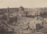 The image displays a historic cityscape with an elevated viewpoint, possibly from a rooftop or higher vantage point. The horizon is seen at the upper part of the composition, showing a transition from the urban environment to rolling hills in the distance. The skyline on the horizon has variations in elevation with a large domed structure at the center and a smaller minaret to its left. The image features a monochromatic palette emphasizing stone and earthy tones of the historic buildings. The urban area near the viewer is filled with stone buildings and ruins, some of which are partially collapsed. Towards the distant hills, there are scattered trees leading to the central dome and surrounding historical architecture.