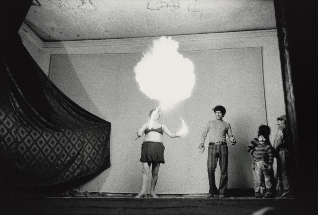 This is a photograph of a performer on a stage creating a fireball. The scene captures a moment of dramatic performance, where a performer in minimal costume stands on a stage with an outstretched arm, holding items creating fire effects, resulting in a large fireball extending upwards. The setting has simple walls and an ornate fabric hanging to one side. The photograph is in black and white, giving it a timeless and gritty aesthetic. Shadows cast on the wall and other individuals observing the performance enhance the dynamism and intensity of the moment.