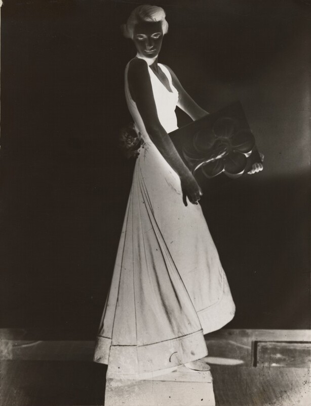 This appears to be a photographic negative which shows a full-body view of a person standing. Their body faces the right, while their head is turned to look over their shoulder towards us. The person is wearing a sleeveless dress that flares out towards the bottom. Their hair is styled back. They are holding a rectangular object with a pattern on it that looks like a large flower. There also appears to be a large flower attached to their dress at the hip. The background, their skin, and the object they hold are all dark gray, while their hair, facial features, and dress are bright white.