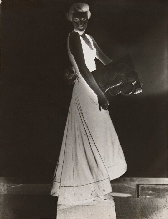 This appears to be a photographic negative which shows a full-body view of a person standing. Their body faces the right, while their head is turned to look over their shoulder towards us. The person is wearing a sleeveless dress that flares out towards the bottom. Their hair is styled back. They are holding a rectangular object with a pattern on it that looks like a large flower. There also appears to be a large flower attached to their dress at the hip. The background, their skin, and the object they hold are all dark gray, while their hair, facial features, and dress are bright white.
