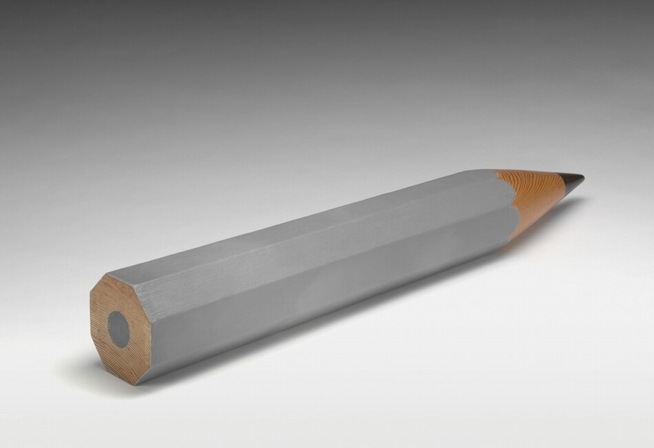 An iron-gray pencil with the stubby proportions of a golf pencil lies with the point angled away from us to our right in this photograph. The other end of the pencil appears cut so the lead is visible. The background lightens from nickel gray to white behind the sculpture.