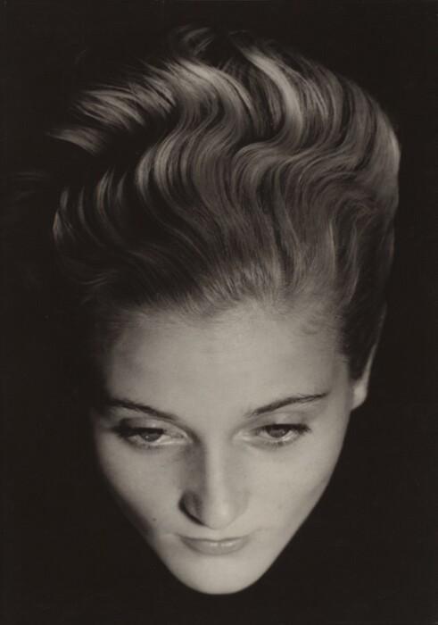 This image depicts a close-up view of a woman's face from above, emphasizing her facial features and hair. Her head is tilted slightly downward, with her eyes looking downwards. Her features include soft lips and gently arched eyebrows. Her hair is styled in elegant waves, adding texture and volume. There are no visible accessories or objects in the background.