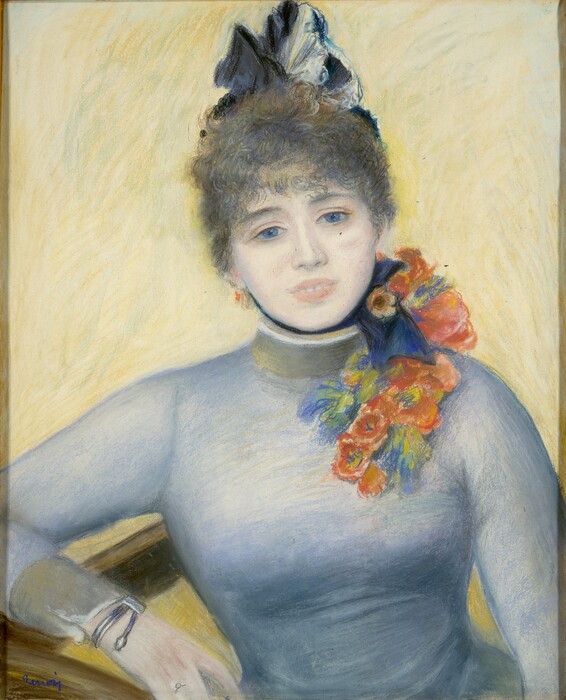 Shown from the waist up in front a yellow background sits a pale-skinned woman wearing a high-necked, long-sleeved blue dress in this vertical pastel. The woman leans slightly to our left as that elbow rests on the arm of a chair or other piece of furniture. She has large blue eyes under dark brows, a round face, and her pink lips are parted to reveal a row of white teeth. Brown tousled curls create bangs across her forehead, and her hair is pulled back under a black and white cap perched on the back of her head. A blue-black ribbon ties in a bow to the right under her chin. Coral-peach, yellow, and pale green flowers cascade from her left shoulder, to our right, down to the chest of her light blue, long-sleeved bodice. The cuff we see is shaded green, and she wears a bracelet and ring on that wrist and hand. Her other arm appears to reach slightly forward and presumably rest in her lap. The woman and the bright yellow background are created with blended strokes, giving the pastel a soft look. The artist signed the lower left corner in cobalt blue, “Renoir.”