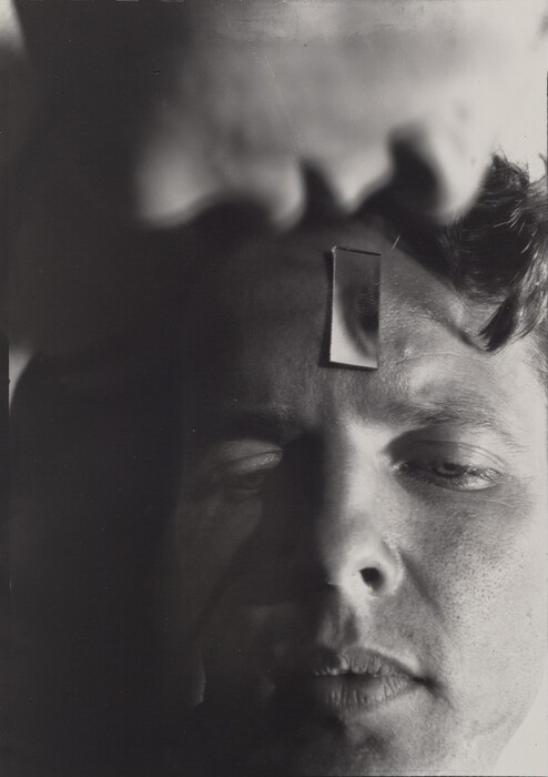 The image shows person's face, with focus on the forehead and eyes. The person's head is slightly tilted to the right. Their facial features include closed or slightly squinted eyes, a prominent forehead, and slightly parted lips. The person has short, curly hair. A small mirror or reflective object is placed vertically on the forehead, showing a reflection of a thumb or finger above the head. The background is out of focus with a blurred light pattern. No clothing, jewelry, or other objects are visible.
