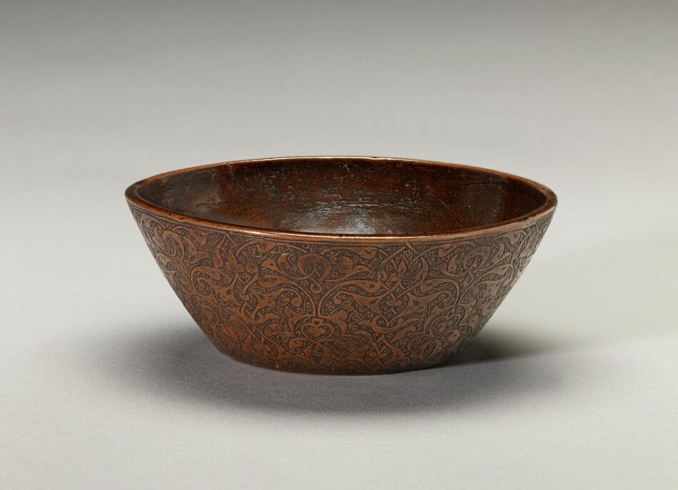 The image displays a sculpted bowl with a circular form. The bowl is a dark metallic brown color, suggesting it is made of metal such as bronze or copper. It is adorned with intricate engraved patterns consisting of interwoven swirling lines and curves. The bowl appears slightly worn with variations in patina but maintains a polished sheen. The angled placement of the bowl in the image showcases both the inner and outer surfaces, emphasizing the craftsmanship of the patterns and the smooth contours of the bowl.