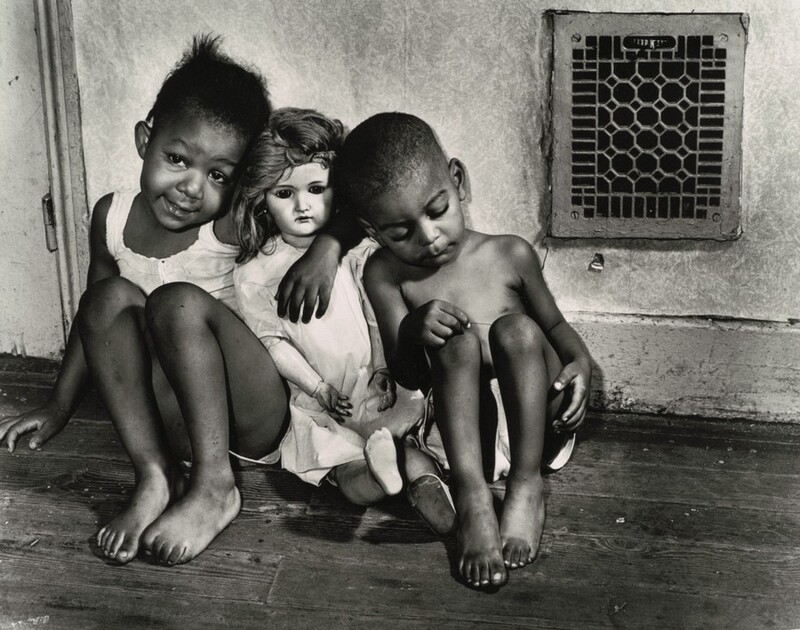 Two Black children sit to either side of a white-skinned doll in this horizontal black and white photograph. The children sit with their knees pulled up on a wood floor against a wall with a grate in the upper right corner of the photograph. A girl sits to our left with one arm around the doll’s shoulders. The girl smiles her head tipped toward the doll, and she curls the toes of one foot. She wears a white, sleeveless top and shorts. The doll is about the same size as the children and wears a white dress and one shoe. The boy wears white shorts and sits to our right, leaning against the doll. He looks down and picks at one knee. The pale-colored wall and high baseboard are scuffed, and the grate is almost square with a geometric design.