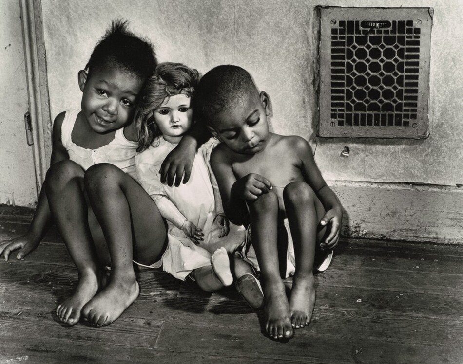 Two Black children sit to either side of a white-skinned doll in this horizontal black and white photograph. The children sit with their knees pulled up on a wood floor against a wall with a grate in the upper right corner of the photograph. A girl sits to our left with one arm around the doll’s shoulders. The girl smiles her head tipped toward the doll, and she curls the toes of one foot. She wears a white, sleeveless top and shorts. The doll is about the same size as the children and wears a white dress and one shoe. The boy wears white shorts and sits to our right, leaning against the doll. He looks down and picks at one knee. The pale-colored wall and high baseboard are scuffed, and the grate is almost square with a geometric design.