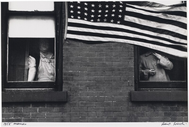 Two people look out of side-by-side windows in a brick building in this horizontal black and white photograph. The woman on our left has a double chin and a broad chest. She wears a polka-dotted dress and dark shawl or vest, and she leans forward with one hand on the sill. Her face is partially shadowed by a white window blind. An American flag hanging near this window billows across the right two-thirds of the picture, entirely hiding the second person’s head. That person wears a coat buttoned up to a rounded collar. They hold one hand near their chin and the other by their waist. The flag has eight columns of stars. The work is signed and dated in the white margin below: “1955 HOBOKEN” to the left and “Robert Frank” to the right.