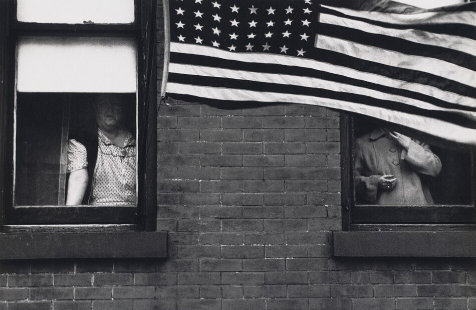 Two people look out of side-by-side windows in a brick building in this horizontal black and white photograph. The woman on our left has a double chin and a broad chest. She wears a polka-dotted dress and dark shawl or vest, and she leans forward with one hand on the sill. Her face is partially shadowed by a white window blind. An American flag hanging near this window billows across the right two-thirds of the picture, entirely hiding the second person’s head. That person wears a coat buttoned up to a rounded collar. They hold one hand near their chin and the other by their waist. The flag has eight columns of stars. The work is signed and dated in the white margin below: “1955 HOBOKEN” to the left and “Robert Frank” to the right.