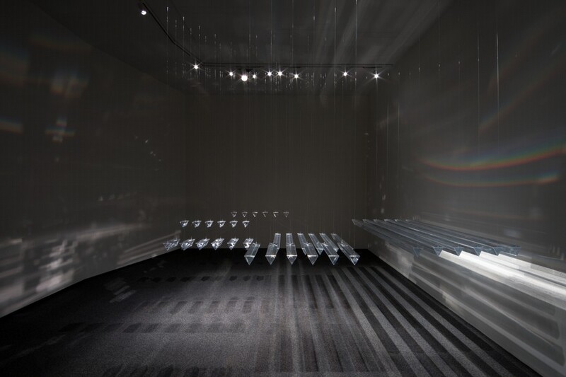 Long prisms hang in rows within a nickel-gray room in this installation. The prisms come toward us in this view. A row of long prisms hang closely together to our right, and shorter prisms are hung in groups farther away from us. They cast long silvery-gray and black shadows on the ground, and hints of rainbows stripe the walls.