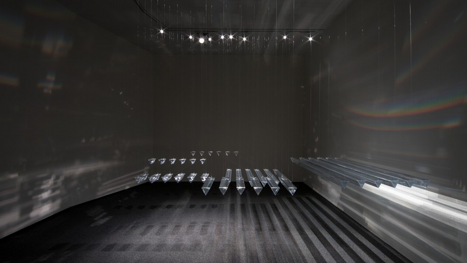 Long prisms hang in rows within a nickel-gray room in this installation. The prisms come toward us in this view. A row of long prisms hang closely together to our right, and shorter prisms are hung in groups farther away from us. They cast long silvery-gray and black shadows on the ground, and hints of rainbows stripe the walls.