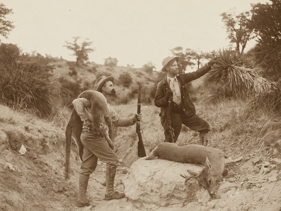 Two men stand outdoors. One man on the left carries a deer over his shoulders, dressed in a patterned shirt, trousers, and boots. He holds a rifle and wears a hat. The man on the right points into the distance and holds a rifle, dressed in a suit and hat. The outdoor setting includes natural vegetation, rocks, and trees in the background.