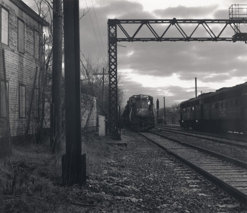 The image displays a railway scene with tracks extending into the distance. A row of trees and industrial structures can be seen near the horizon, against a cloudy sky. In the foreground, there is a weathered building, wire fence, power lines, and utility poles. Two trains are visible: one stationary, labeled "Conrail," and another moving on the opposite side. The color scheme is monochromatic with various shades of gray, and the lighting is subdued due to overcast clouds.
