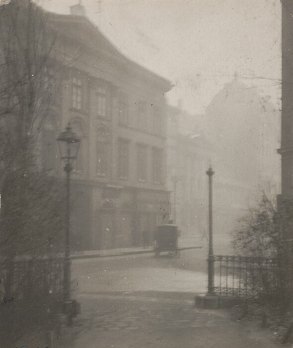 The image shows a street scene featuring a cobblestone path in an urban setting. The view is from ground level, looking down the path. The scene is captured in monochrome, with shades of grey dominating. A building with arched windows is on the left, fading into mist in the distance. In the center, there is a streetlamp and a faintly visible cart. On the right, there is an ornate metal railing and bare trees. The image lacks brushstroke details, suggesting it is a photograph. Objects close to the viewer transition into blurred contours of buildings and streets disappearing into the mist.