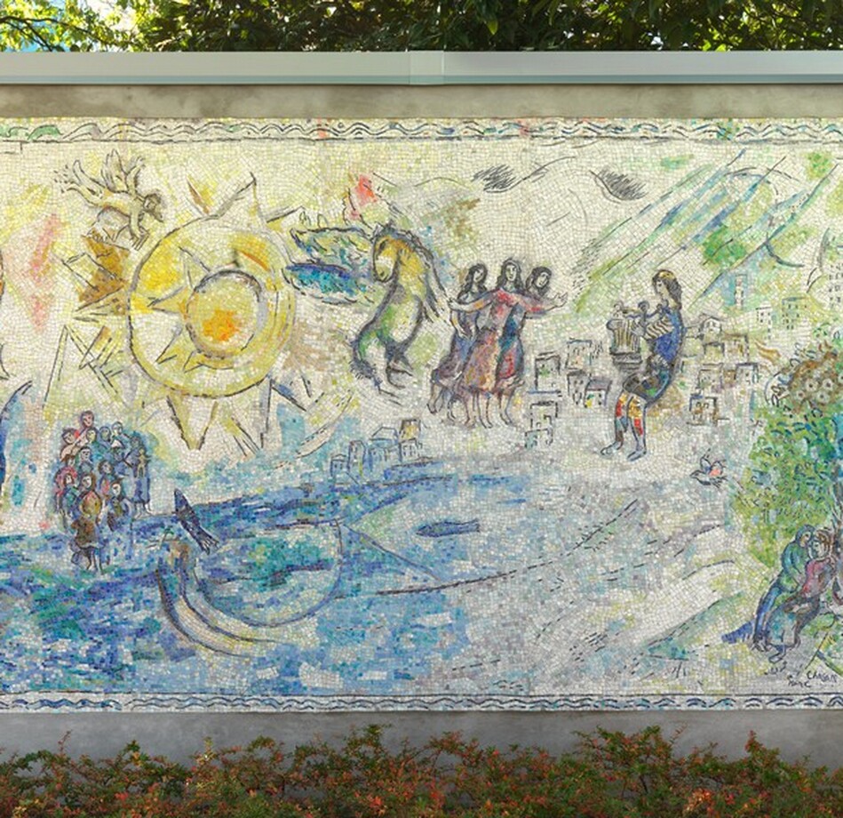 Created with tiny, colored stones and glass, this mosaic shows groups of people along and near the edge of a brilliant, topaz-blue body of water with a large, lemon-yellow, stylized sun in the sky in this horizontal composition. The aquamarine blue and vibrant yellow along with lime green, lilac purple, oyster white, and teal used in the people and landscape give this work a shimmering look. A pair of people recline together under a tree in the lower right corner. A sheep stands nearby, and a bird perches in the tree above. Beyond the tree and to our left, a cluster of box-like forms suggests a city in the distance. Larger in scale, so seeming closer to us, a man wearing a lapis-blue toga and a yellow cap holds a stringed instrument, a lyre, in front of his body. To the left, at the center of the composition, a trio of women each wearing amethyst-purple, magenta-pink, or lapis-blue dresses stand close to each other. They have long, dark hair and their bodies and garments are outlined in black. Between them and the large sun to our left, a winged horse with a buttercup-yellow body and turquoise-blue wings rears up. Near the upper left corner, the stylized sun is represented by a disk surrounded by pointed peaks, surrounded by a larger disk outlined with another ring of triangular points. A winged person holding a set of pan pipes flies above the sun to our left, over a small crescent moon below. Near the lower left corner, two groups of people gather around a body of bright blue water occupied by two fish. The artist signed and dated the work in the lower right corner: “MArC ChAgAll 69.” The mosaic is displayed outside in front of a screen of trees, with a band of bushes with green and orange leaves below.