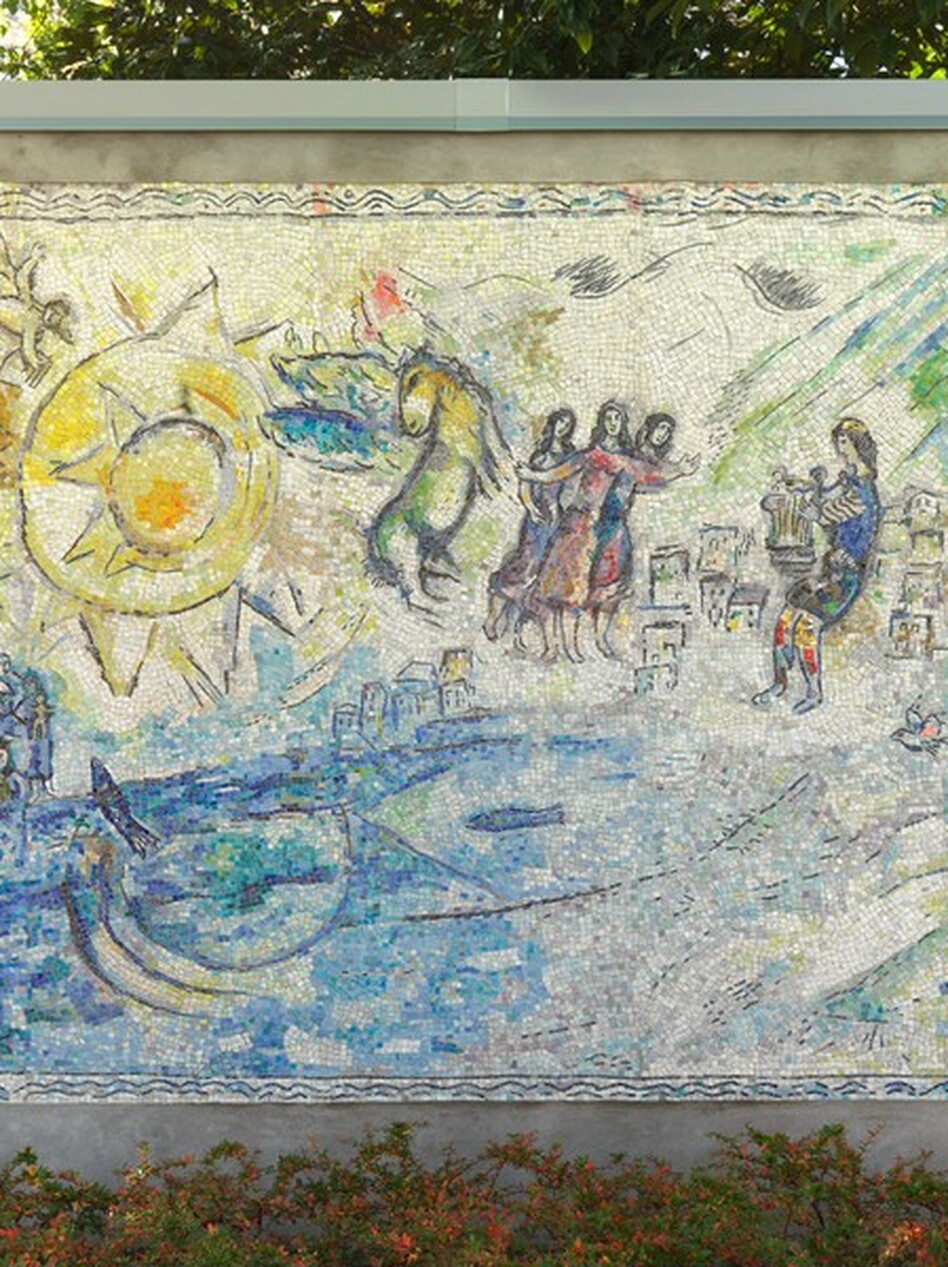Created with tiny, colored stones and glass, this mosaic shows groups of people along and near the edge of a brilliant, topaz-blue body of water with a large, lemon-yellow, stylized sun in the sky in this horizontal composition. The aquamarine blue and vibrant yellow along with lime green, lilac purple, oyster white, and teal used in the people and landscape give this work a shimmering look. A pair of people recline together under a tree in the lower right corner. A sheep stands nearby, and a bird perches in the tree above. Beyond the tree and to our left, a cluster of box-like forms suggests a city in the distance. Larger in scale, so seeming closer to us, a man wearing a lapis-blue toga and a yellow cap holds a stringed instrument, a lyre, in front of his body. To the left, at the center of the composition, a trio of women each wearing amethyst-purple, magenta-pink, or lapis-blue dresses stand close to each other. They have long, dark hair and their bodies and garments are outlined in black. Between them and the large sun to our left, a winged horse with a buttercup-yellow body and turquoise-blue wings rears up. Near the upper left corner, the stylized sun is represented by a disk surrounded by pointed peaks, surrounded by a larger disk outlined with another ring of triangular points. A winged person holding a set of pan pipes flies above the sun to our left, over a small crescent moon below. Near the lower left corner, two groups of people gather around a body of bright blue water occupied by two fish. The artist signed and dated the work in the lower right corner: “MArC ChAgAll 69.” The mosaic is displayed outside in front of a screen of trees, with a band of bushes with green and orange leaves below.