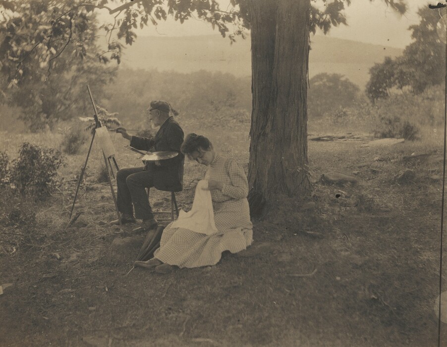 A man sits painting at an easel set up under a tree and a woman sits nearby, her back to the trunk, sewing a white cloth in this horizontal black and white photograph. The tree reaches off the top edge of the composition to our right of center. The two people sit on the left half, angled in that direction so we see them almost in profile. The man wears a floppy cap and dark suit. He holds an oval paint palette in one hand and lifts a paintbrush to the painting with the other. The woman’s dark hair is gathered atop her head, and she looks down at her work. She wears a long-sleeved, ankle-length dress with a high collar. Bushes and mountains are hazy in the distance.