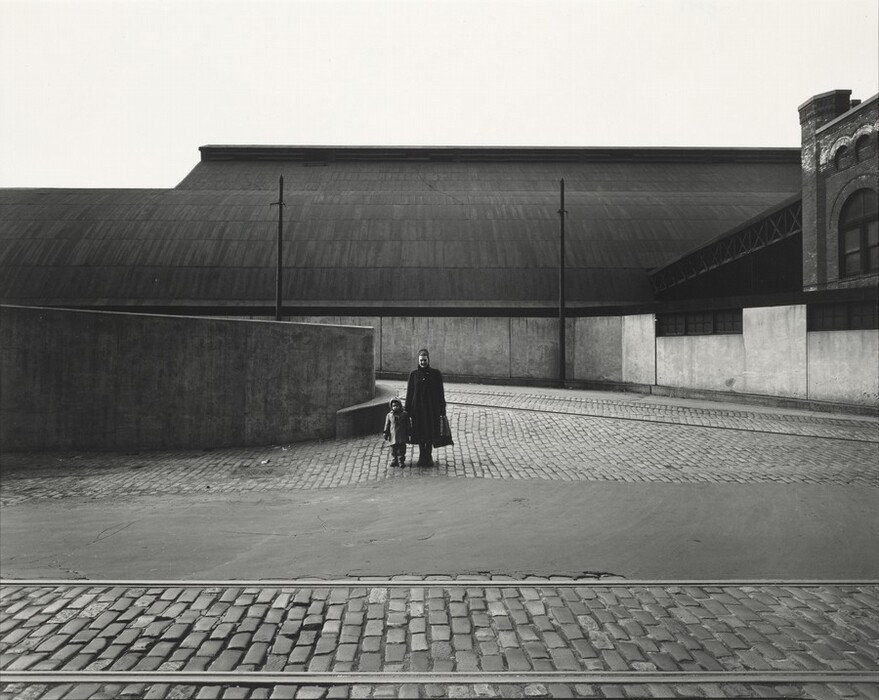 The image shows a woman and a child standing on a cobblestone street. The woman, on the right, has short, dark hair and wears a long dark coat. She is holding the child's hand. The child wears a cap and a coat and stands close to the woman. In the background, there is a large industrial building with a curved roof and large walls. The street surfaces are wet, and tram tracks are visible. The setting has a stark, industrial appearance with minimal adornment, suggesting an urban environment.