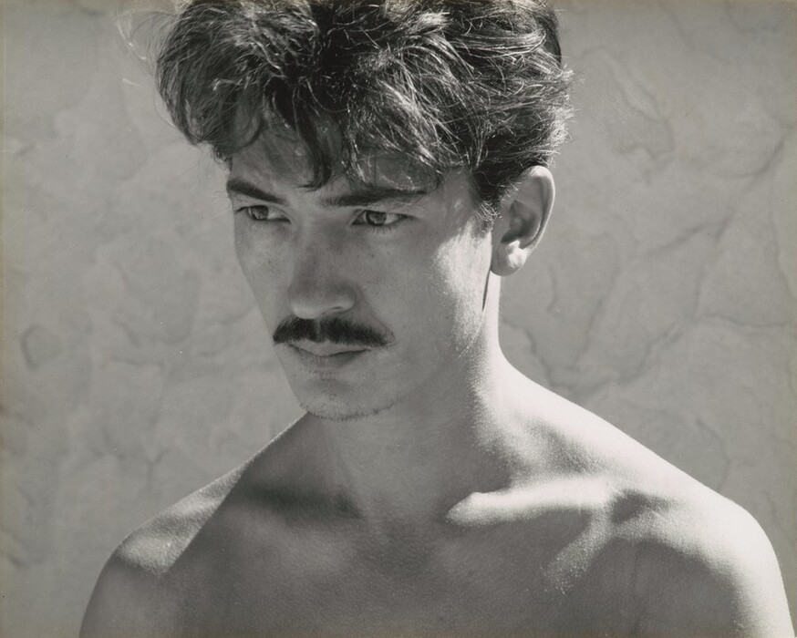 The image shows a person in a close-up, with their upper body visible, including their shoulders. The individual is facing slightly to the side, looking off into the distance with a neutral expression. They have a defined mustache and prominent cheekbones. Their dark hair is styled in a slightly tousled manner. The person is not wearing a shirt, and no other clothing is visible. There are no visible accessories or objects in their hands. The background appears to be a textured surface, likely a wall, with various shades suggesting light and shadow play. The overall tone of the image is monochromatic.
