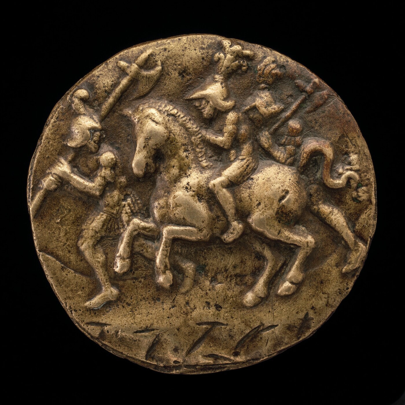The sculpture depicted on the round object shows a mounted warrior and an infantry soldier. The central figure is a man riding a horse, wearing a helmet with a crest and holding a spear or staff. The horse is depicted mid-stride. In front of the horse, there is an infantry soldier wearing a helmet and holding a spear or staff. The engraving is on a raised, circular surface, made of a gold-colored metal. The texture appears rough and aged, with skillfully carved details.