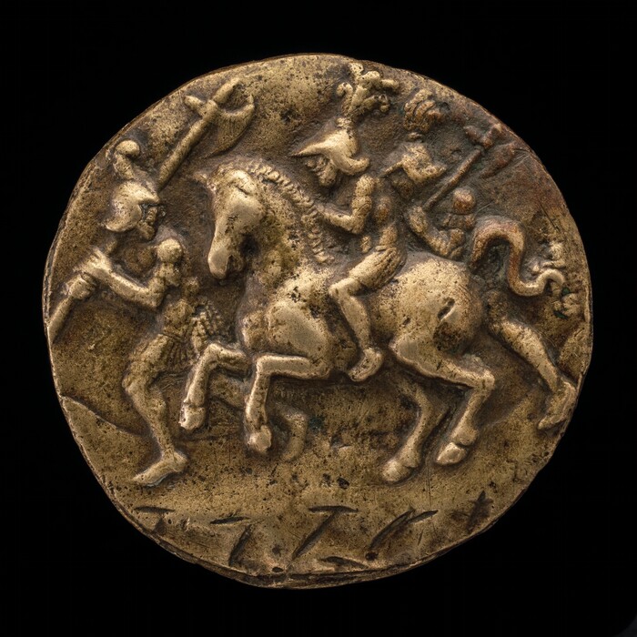 The sculpture depicted on the round object shows a mounted warrior and an infantry soldier. The central figure is a man riding a horse, wearing a helmet with a crest and holding a spear or staff. The horse is depicted mid-stride. In front of the horse, there is an infantry soldier wearing a helmet and holding a spear or staff. The engraving is on a raised, circular surface, made of a gold-colored metal. The texture appears rough and aged, with skillfully carved details.