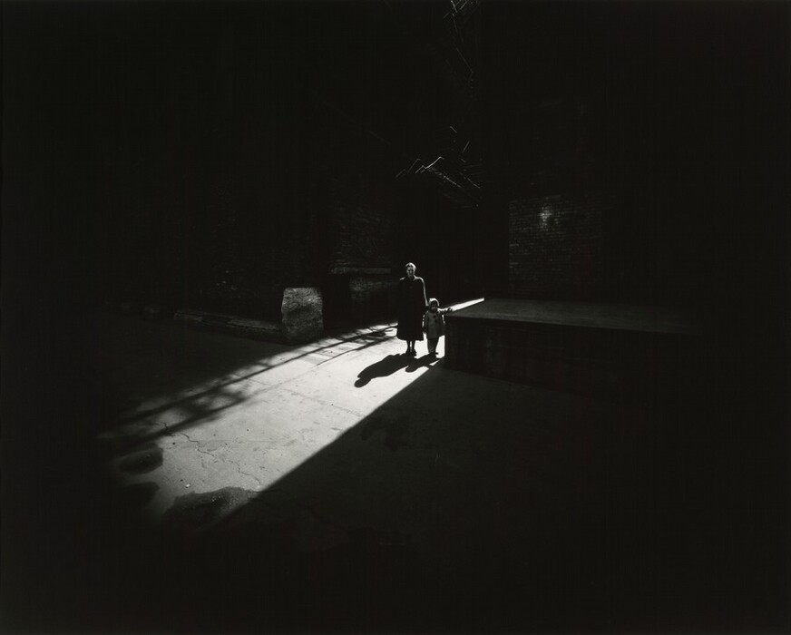 The image shows two people, one adult and one child, facing the camera in dramatic lighting. The adult wears a dark coat, while the child wears a light-colored coat and a hat. They stand in a brightly lit area with dark surroundings, against large brick walls and concrete surfaces.