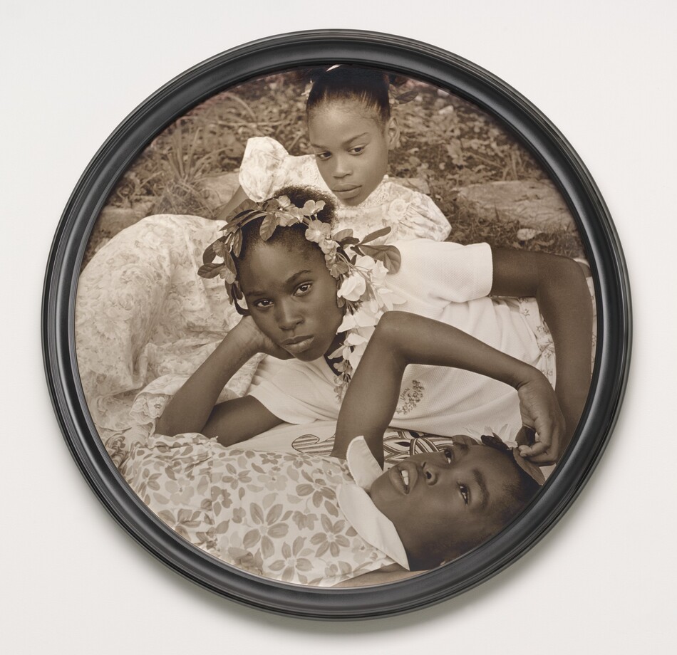 Three young Black girls lie on the grass in this closely cropped, sepia-toned, circular photograph so their faces roughly line up near the center. At the bottom of the composition, one girl lies on her back and looks up into the sky. Her head, torso, and right arm are visible. She wears a floral-patterned dress and holds her right hand up to the top of her head. The second girl reclines on her right side behind the first, so she is angled to our left. She props her head in her right hand and looks steadily at us. Her face hovers at the center of the composition. She wears a white t-shirt and a garland encircles her head. The third girl, at the top of the composition, seems to prop her body up on her left elbow. She wears a floral dress and looks down and to our right. Grass and paving rocks fill the space behind her.