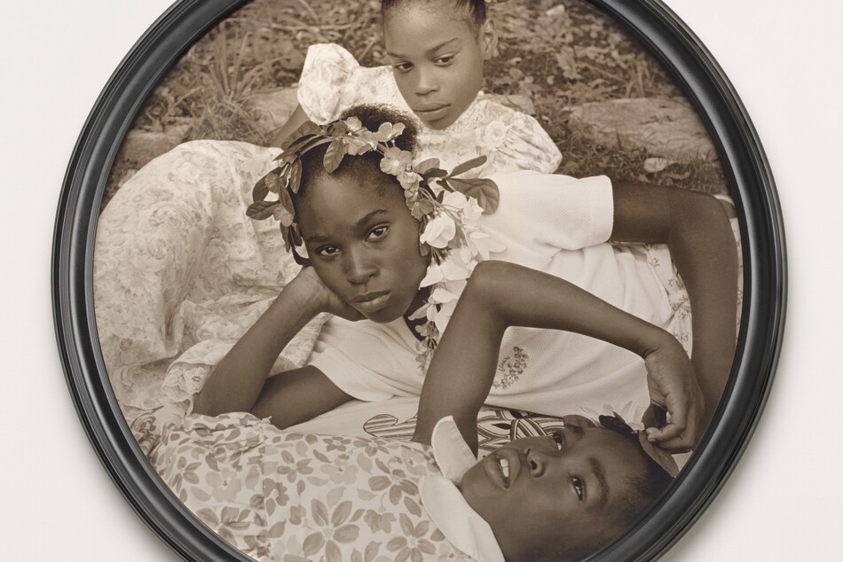 Three young Black girls lie on the grass in this closely cropped, sepia-toned, circular photograph so their faces roughly line up near the center. At the bottom of the composition, one girl lies on her back and looks up into the sky. Her head, torso, and right arm are visible. She wears a floral-patterned dress and holds her right hand up to the top of her head. The second girl reclines on her right side behind the first, so she is angled to our left. She props her head in her right hand and looks steadily at us. Her face hovers at the center of the composition. She wears a white t-shirt and a garland encircles her head. The third girl, at the top of the composition, seems to prop her body up on her left elbow. She wears a floral dress and looks down and to our right. Grass and paving rocks fill the space behind her.