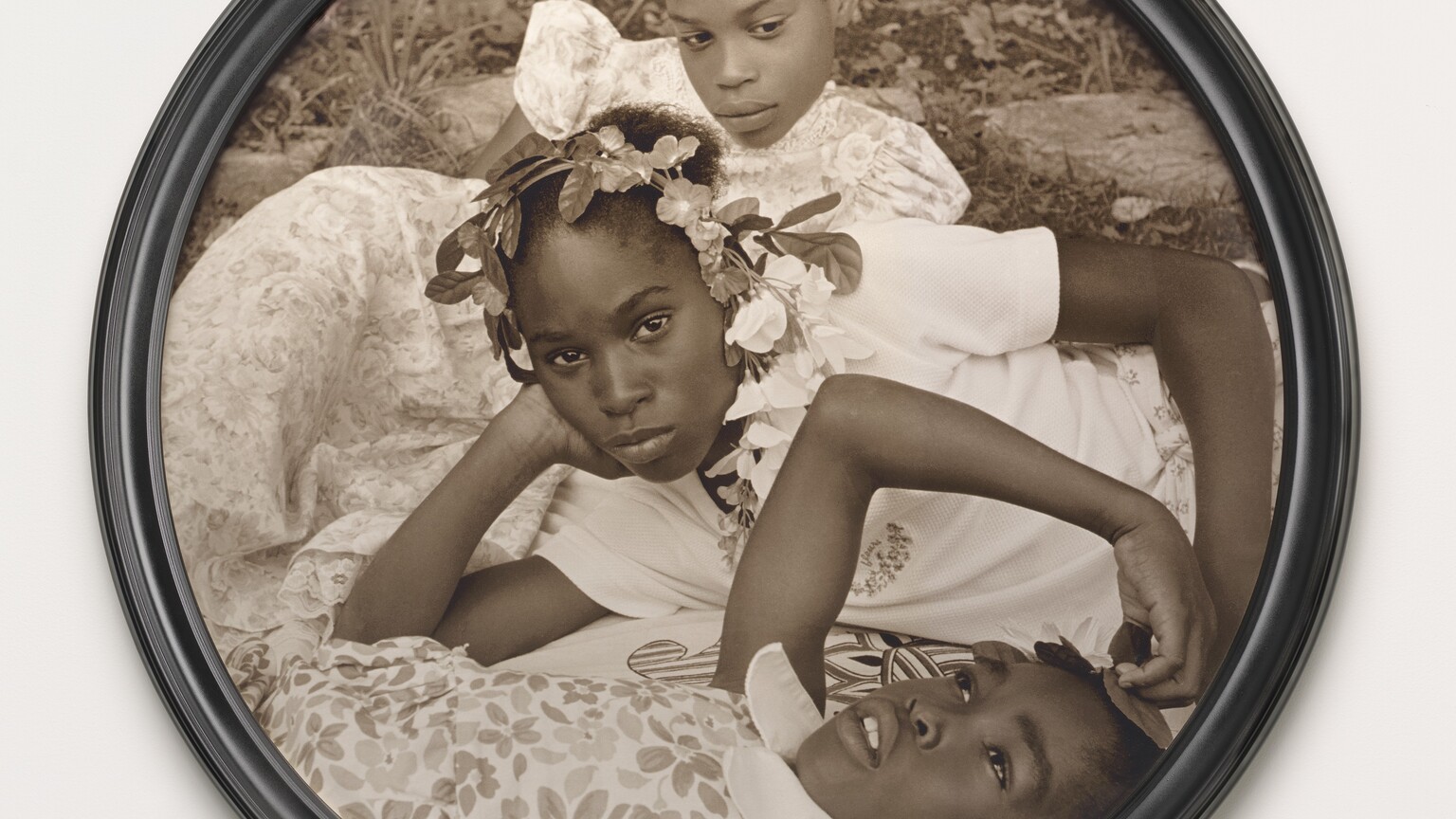 Three young Black girls lie on the grass in this closely cropped, sepia-toned, circular photograph so their faces roughly line up near the center. At the bottom of the composition, one girl lies on her back and looks up into the sky. Her head, torso, and right arm are visible. She wears a floral-patterned dress and holds her right hand up to the top of her head. The second girl reclines on her right side behind the first, so she is angled to our left. She props her head in her right hand and looks steadily at us. Her face hovers at the center of the composition. She wears a white t-shirt and a garland encircles her head. The third girl, at the top of the composition, seems to prop her body up on her left elbow. She wears a floral dress and looks down and to our right. Grass and paving rocks fill the space behind her.