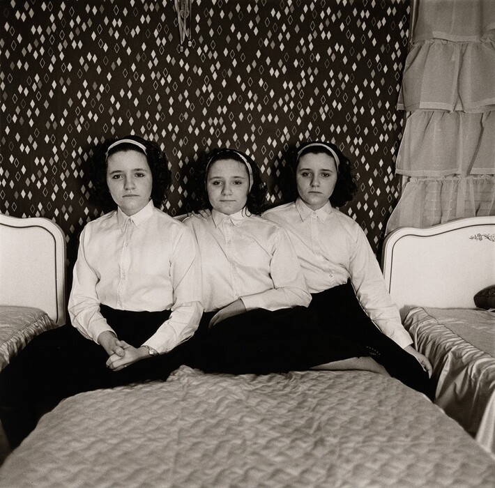 In this black-and-white photograph, three identical young women are seated side by side on a bed in a bedroom. They are positioned closely together, facing the camera. Each has an oval face, pale skin, and wavy dark hair pulled back with a white headband, and they are all wearing white shirts and dark skirts. The woman on the left has her hands clasped in her lap. The room has patterned wallpaper with diamond-like designs on a dark background, and on either side of the central bed, partial views of bed headboards and ruffled curtains are visible.